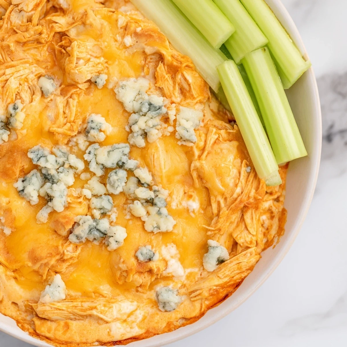 Steaming hot Spicy Buffalo Chicken Dip in a casserole dish, topped with melted cheddar and blue cheese, with crisp celery sticks for dipping.