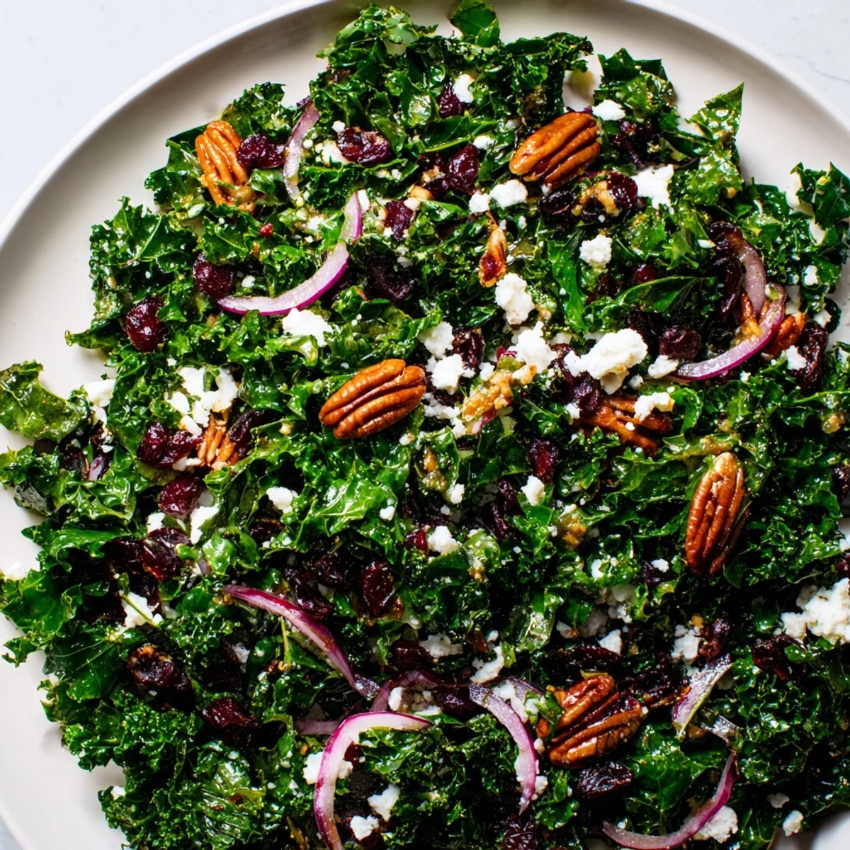 Freshly massaged kale tossed with sweet dried cranberries and crunchy toasted pecans in a tangy maple vinaigrette. The Winter Kale Salad with Pecans and Cranberries is served on a white plate with thin apple slices.