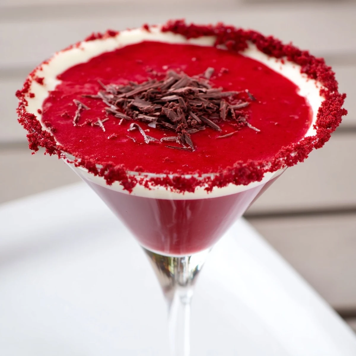 A chilled Decadent Red Velvet Martini rests beside chocolate shavings, showcasing its creamy texture and deep red color against a cozy home bar backdrop.