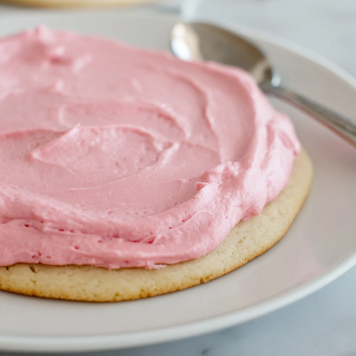 Thick, soft Crumbl Sugar Cookies with creamy pink almond frosting on a white plate, ready to serve at a party.