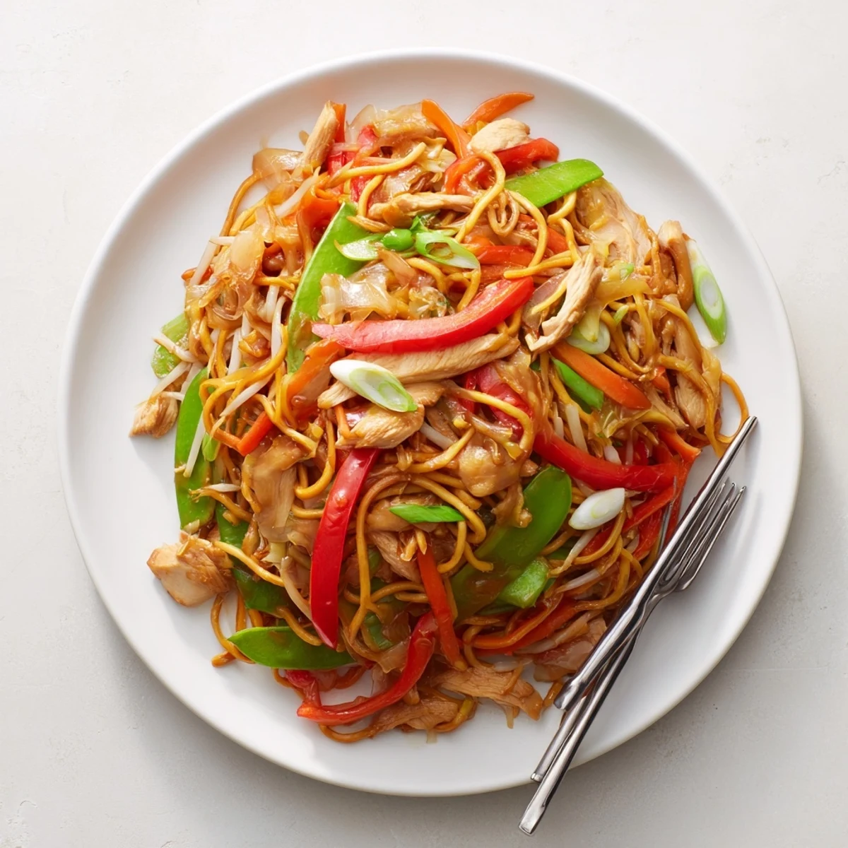 Plate of savory Chicken Chow Mein noodles piled high, garnished with scallions and served alongside a small dish of chili oil.  