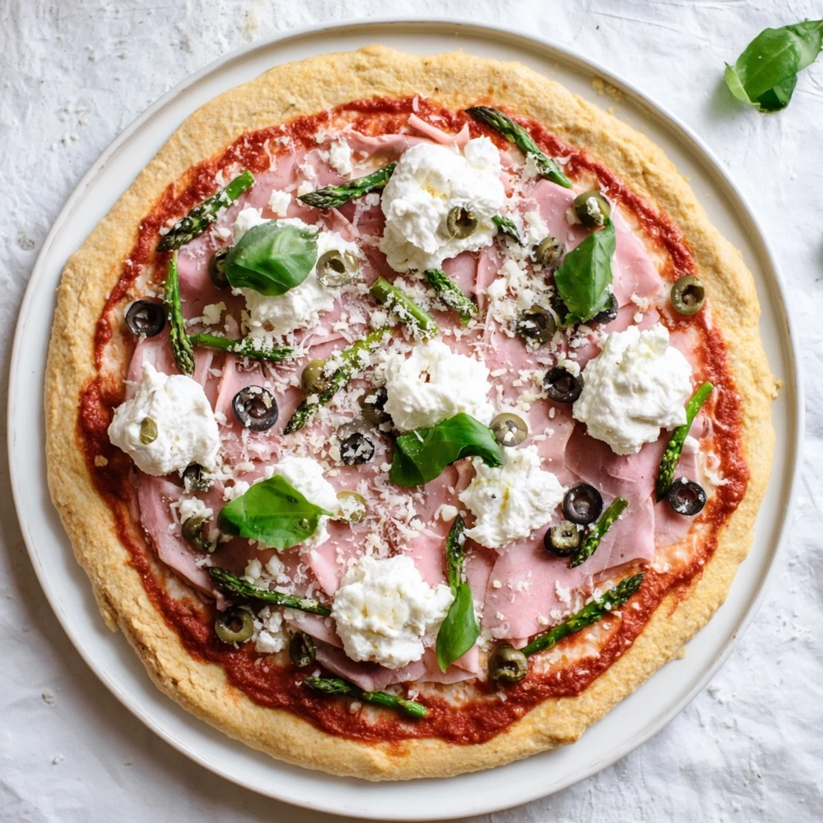 A close-up of a freshly baked Ham, Olive, Asparagus, and Ricotta Pizza on a rustic wooden board, garnished with fresh basil leaves.  