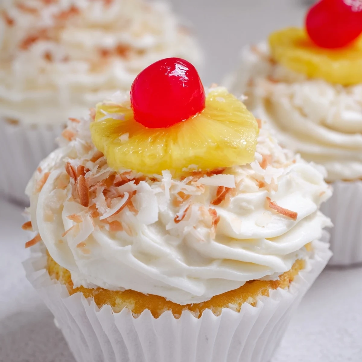 Golden Pina Colada cupcakes topped with fluffy white coconut frosting and garnished with toasted coconut flakes and a cherry