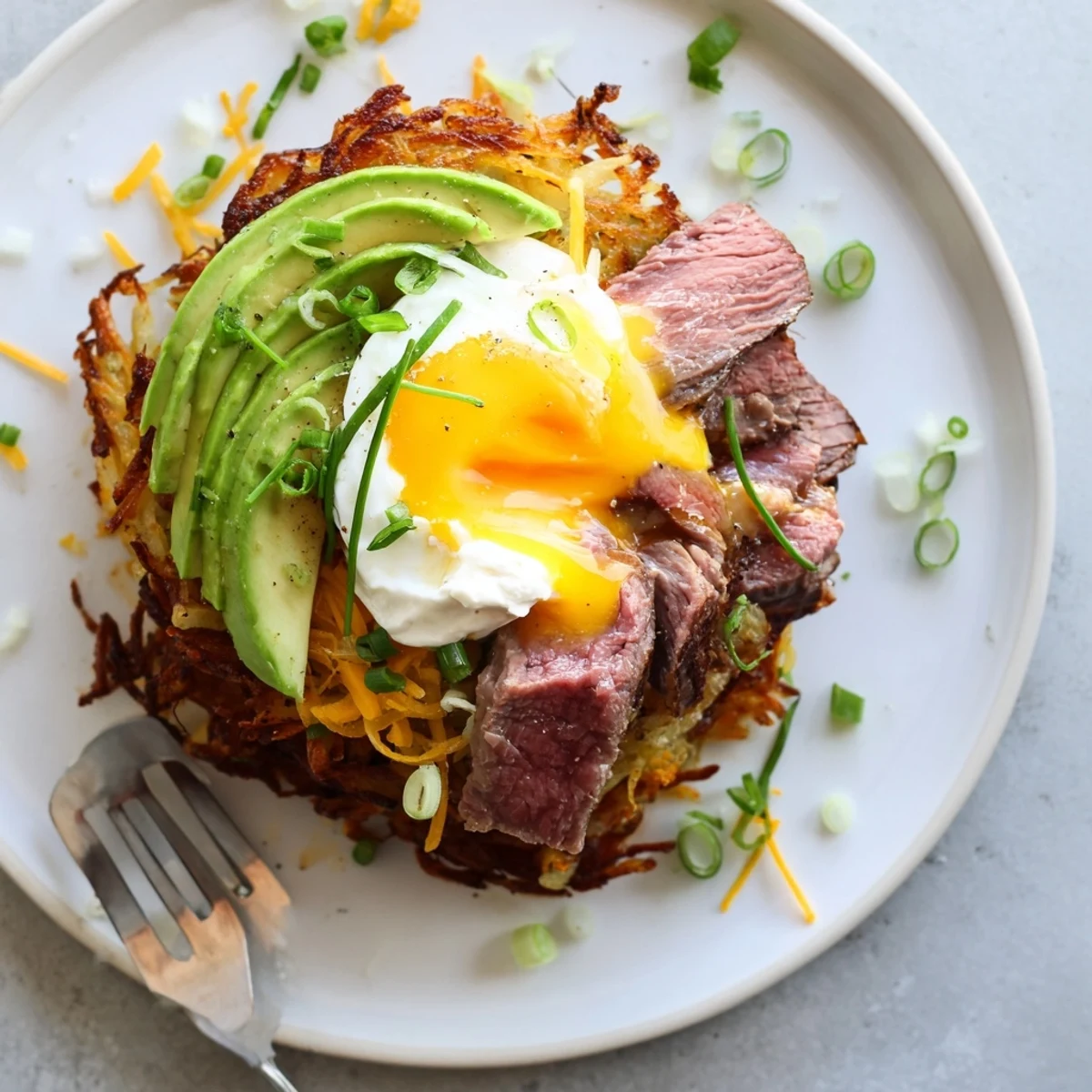 Cast iron-served Loaded Steak Egg Hash Brown Stacks garnished with avocado and chives