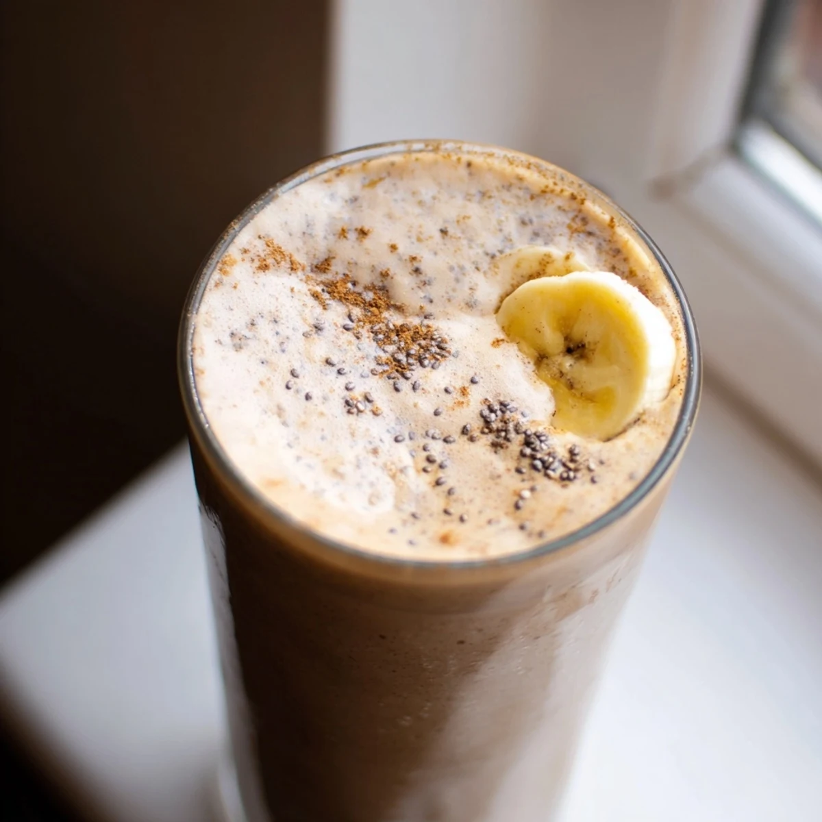 Thick frothy banana protein coffee smoothie topped with a dusting of cinnamon in a tall clear serving glass